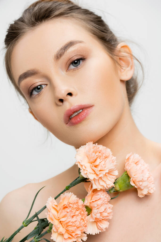портрет молодой и очаровательной женщины с идеальным лицом, смотрящей в камеру рядом со свежими цветами, изолированными на сером - Фото, изображение