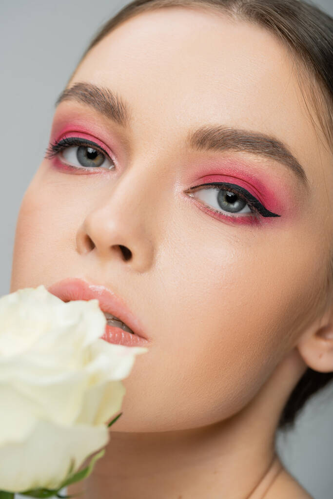 close up portrait of woman with pink eye shadows looking at camera near ivory rose isolated on grey - Foto, Bild