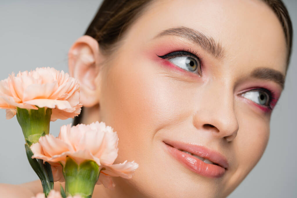 close up portrait of smiling woman with makeup looking away near peach carnations isolated on grey - Foto, Imagem