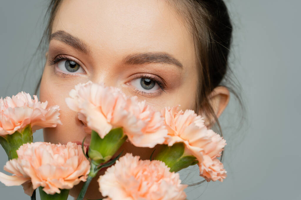 portrait of young woman looking at camera near peach carnations isolated on grey - Foto, afbeelding