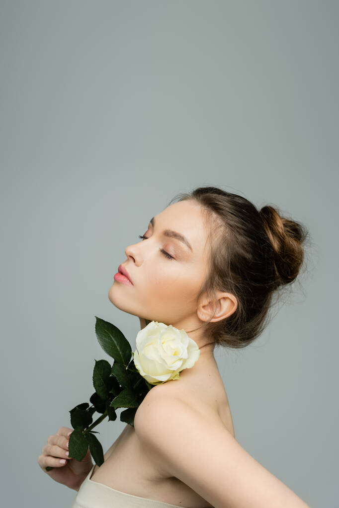 чувственная женщина с закрытыми глазами, держащая свежую розу возле обнаженного плеча, изолированного на сером - Фото, изображение
