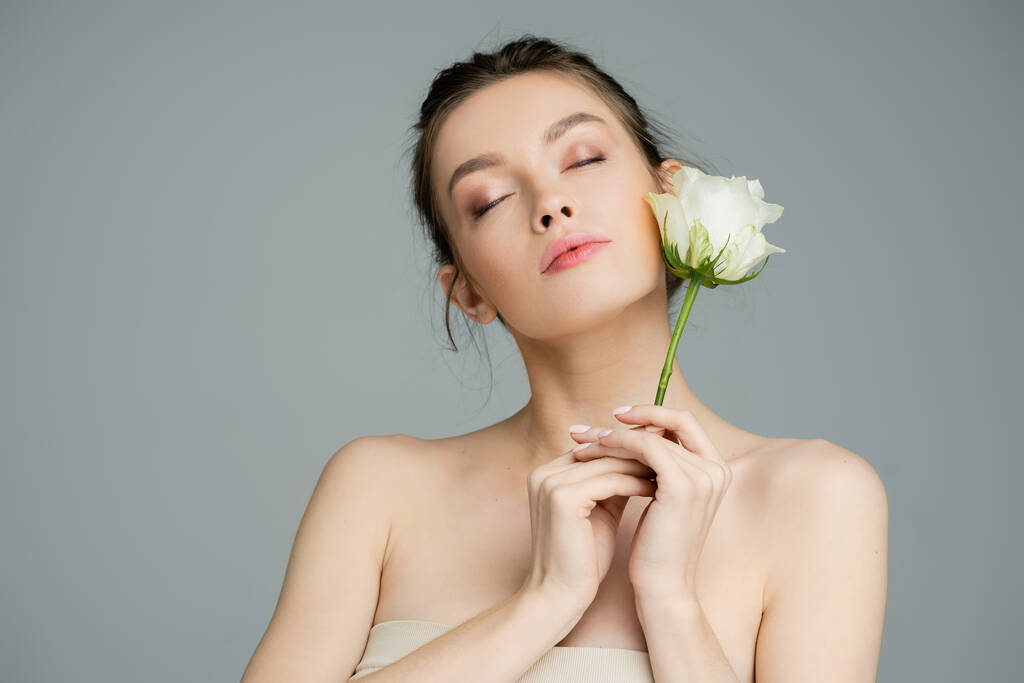 молода жінка з голими плечима і закритими очима тримає білу троянду ізольовано на сірому
 - Фото, зображення