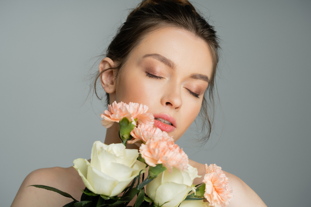 Mujer joven con maquillaje natural sosteniendo flores aisladas en gris  - Foto, imagen