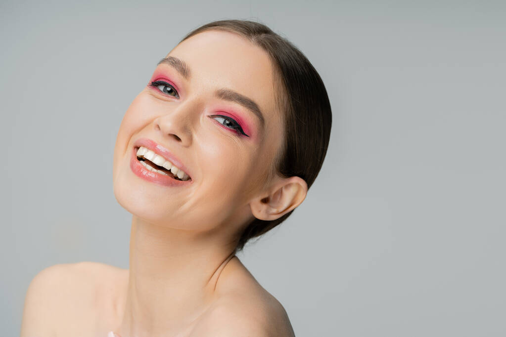 Cheerful woman with naked shoulders and and bright pink makeup looking at camera isolated on grey - Foto, immagini