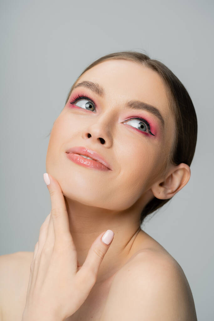 Portrait of joyful woman with pink eye shadow touching face isolated on grey  - Foto, imagen