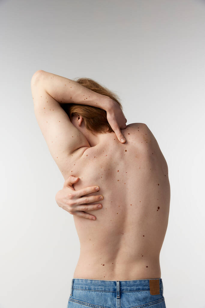 Φωτογραφία του νεαρού άνδρα που ποζάρει γυμνός με τζιν πάνω από γκρι φόντο στούντιο. Τρύπες στο σώμα, υγιής, δυνατή πλάτη, σπονδυλική στήλη. Έννοια της ανδρικής υγείας, της φροντίδας σώματος και δέρματος, της υγιεινής και της ανδρικής κοσμετολογίας - Φωτογραφία, εικόνα