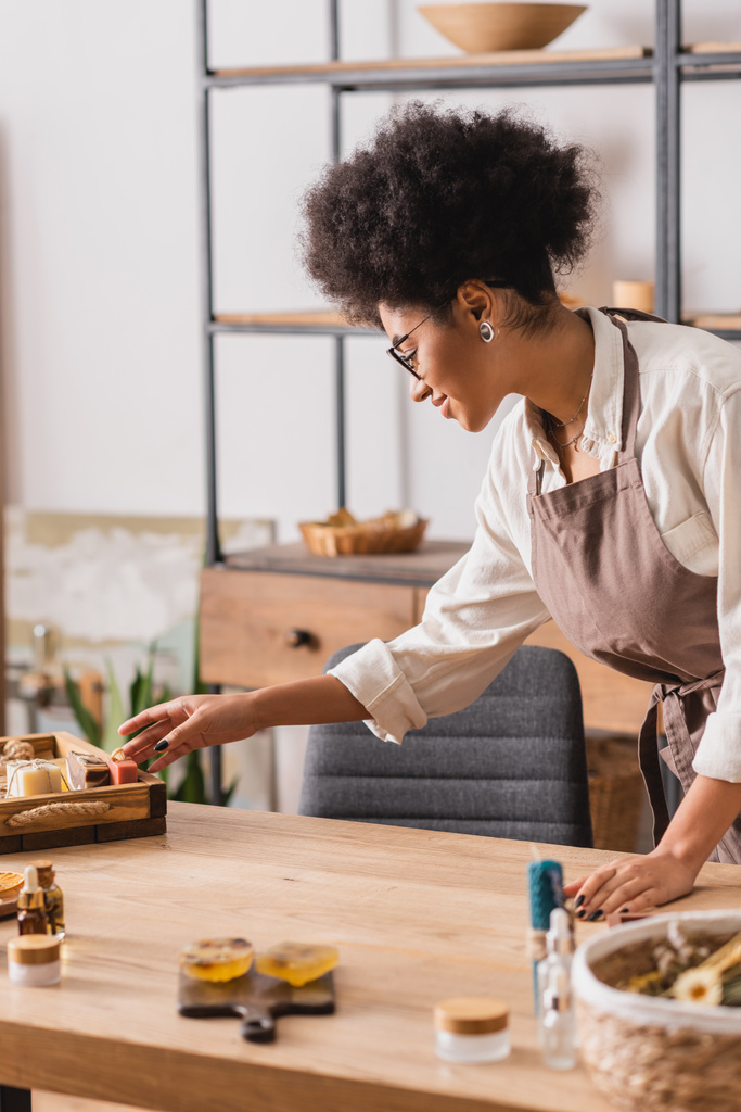 Afrikaans-Amerikaanse vrouw in bril en schort bereiken zeep bars bijna wazig ingrediënten op tafel in ambachtelijke werkplaats - Foto, afbeelding