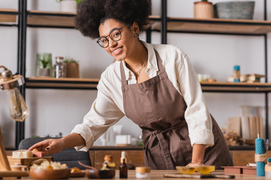 sonriente artesana afroamericana en gafas y delantal llegando a jabón hecho a mano cerca de ingredientes borrosos en la mesa - Foto, imagen