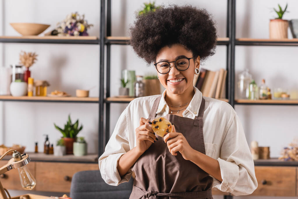 Веселая африканская американка в очках и фартуке держит мыло ручной работы возле стойки с натуральными ингредиентами на размытом фоне - Фото, изображение