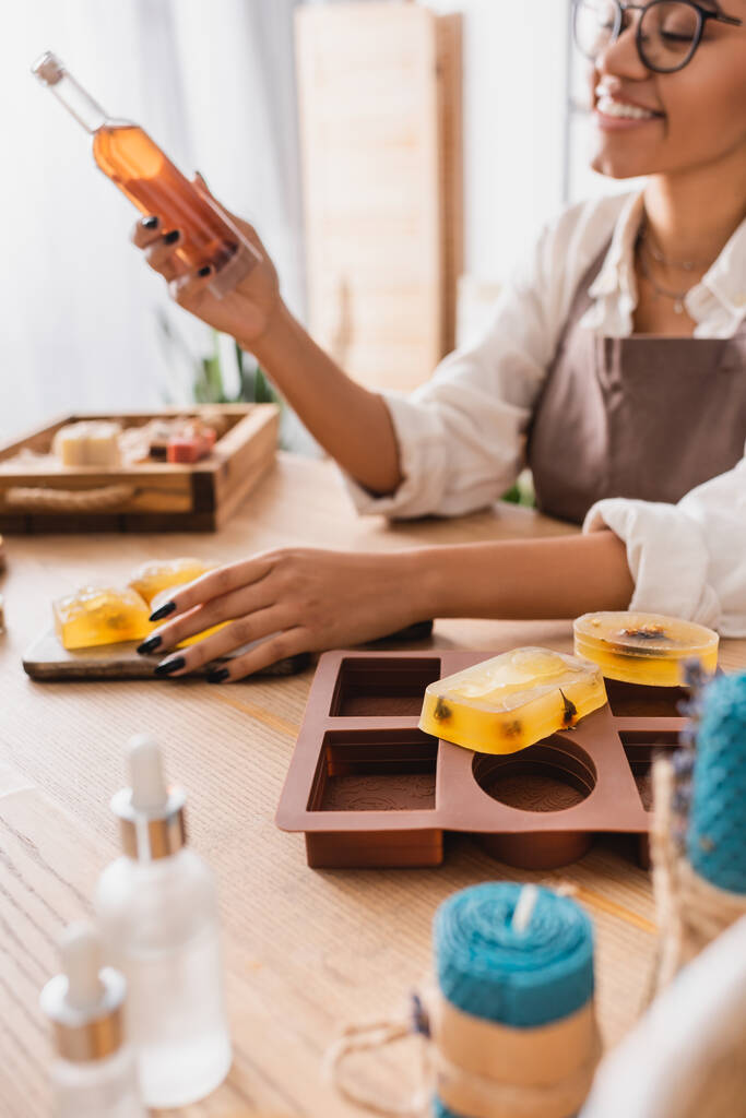 foyer sélectif de savon fait à la main et moule en silicone près heureuse femme afro-américaine avec bouteille d'huile d'olive sur fond flou - Photo, image