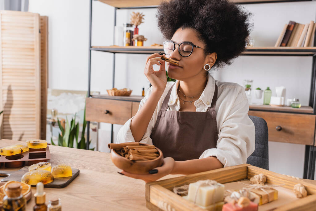 довольная американская ремесленница из Африки в очках наслаждается вкусом корицы рядом с мылом ручной работы в мастерской - Фото, изображение