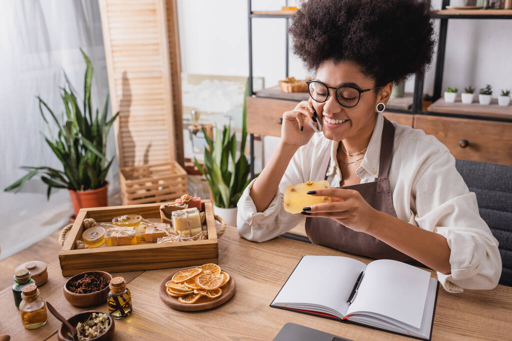 heureuse femme afro-américaine tenant du savon fait maison et parlant sur téléphone portable près d'ingrédients naturels et ordinateur portable - Photo, image