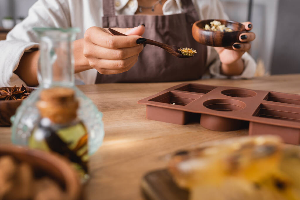 cropped view of african american craftswoman holding wooden bowl and spoon with dried herbs near silicone mold on blurred foreground - Photo, Image