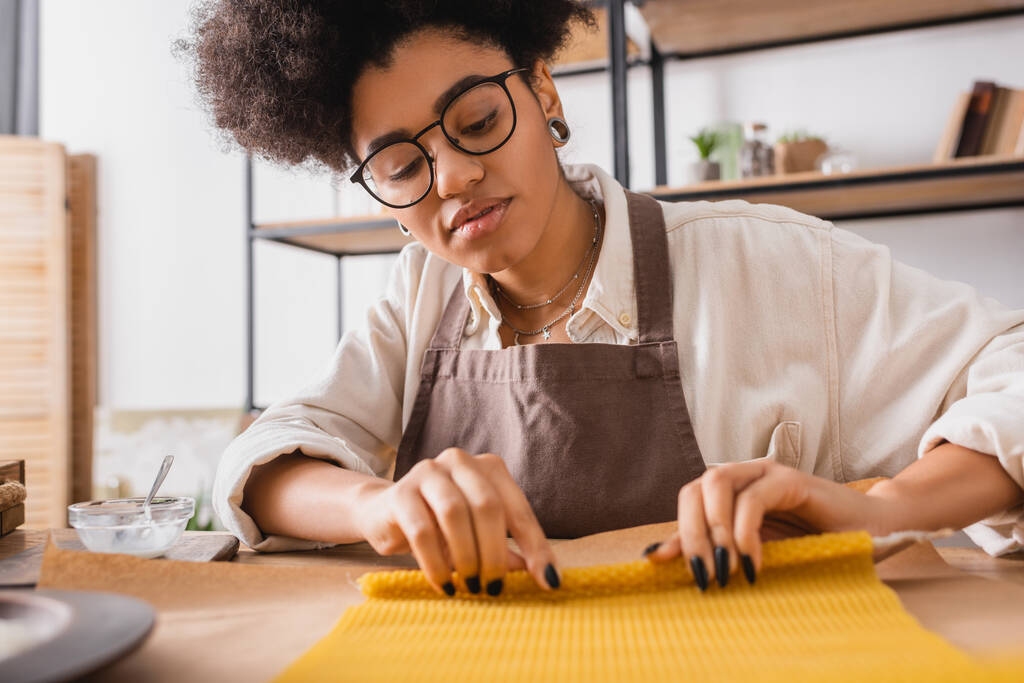 artesana afroamericana en delantal y anteojos rodando hoja de cera en pergamino mientras hace vela en taller - Foto, Imagen