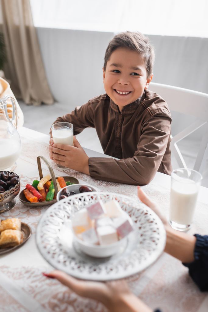 Cheerful muslim boy holding milk and looking at mom holding turkish delight during ramadan  - Fotó, kép