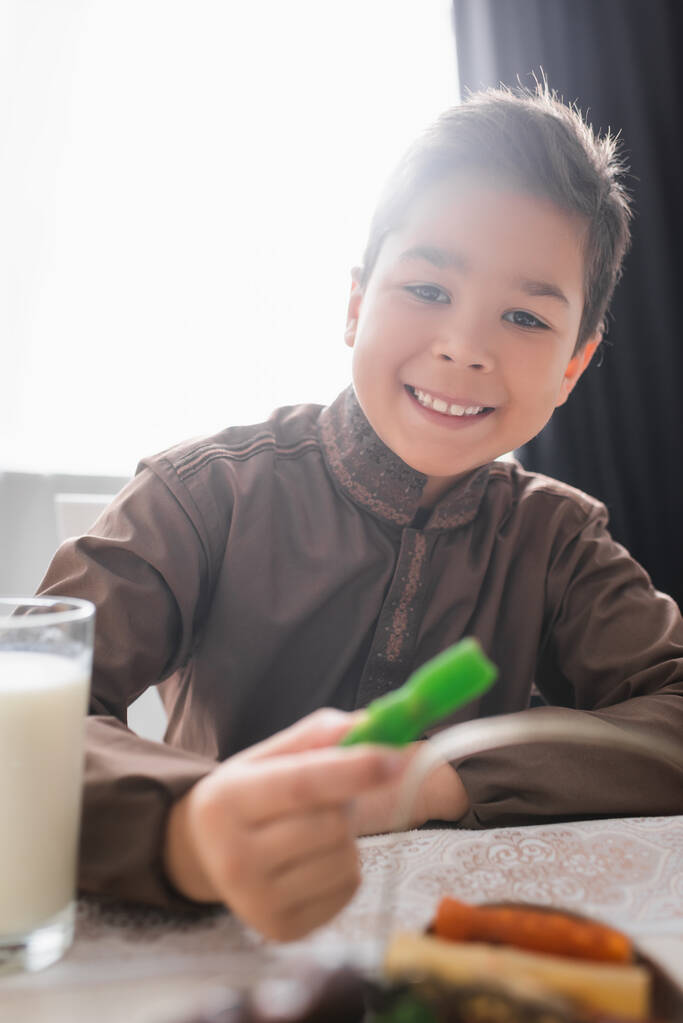 Sonriente niño musulmán sosteniendo borrosa cevizli sucuk cerca de vaso de leche durante el desayuno ramadán  - Foto, Imagen
