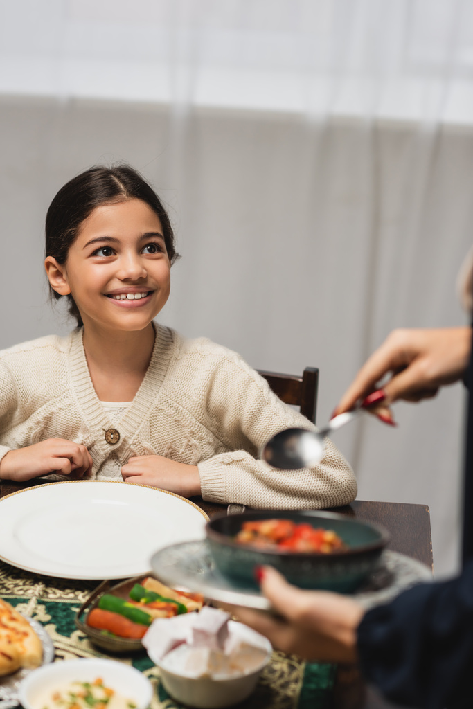 Sonriente chica musulmana mirando a mamá borrosa con comida durante la cena de ramadán en casa  - Foto, Imagen