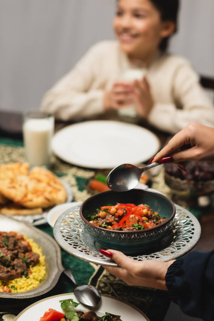 Muslim woman holding tasty dish near blurred daughter and ramadan dinner  - Fotoğraf, Görsel