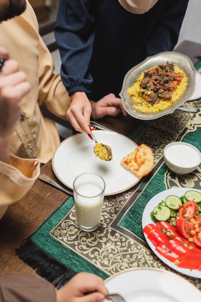 Vista recortada de la mujer musulmana sirviendo pilaf en el plato cerca de la familia durante el ramadán  - Foto, imagen