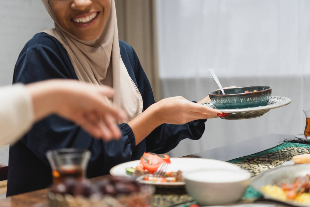 Καλλιεργημένη θέα χαμογελαστή μουσουλμάνα γυναίκα κρατώντας το φαγητό κοντά στην κόρη στο σπίτι  - Φωτογραφία, εικόνα