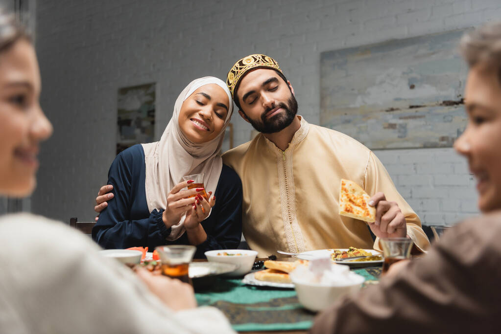 Arabian man hugging wife in hijab near blurred kids during iftar at home  - 写真・画像