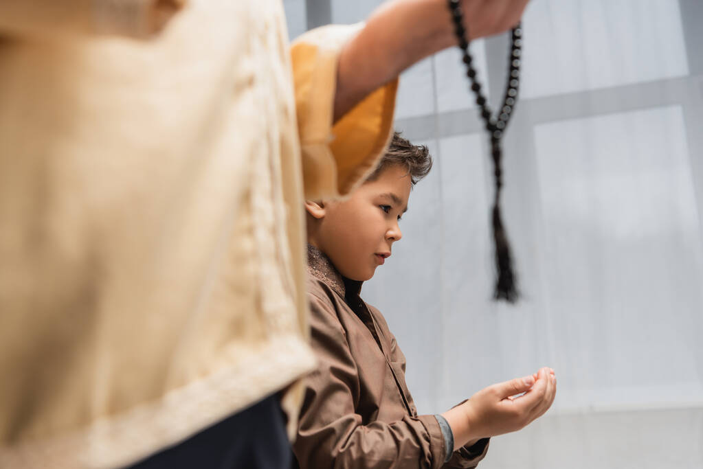 Middle eastern boy praying near blurred dad with prayer beads at home  - Foto, Bild