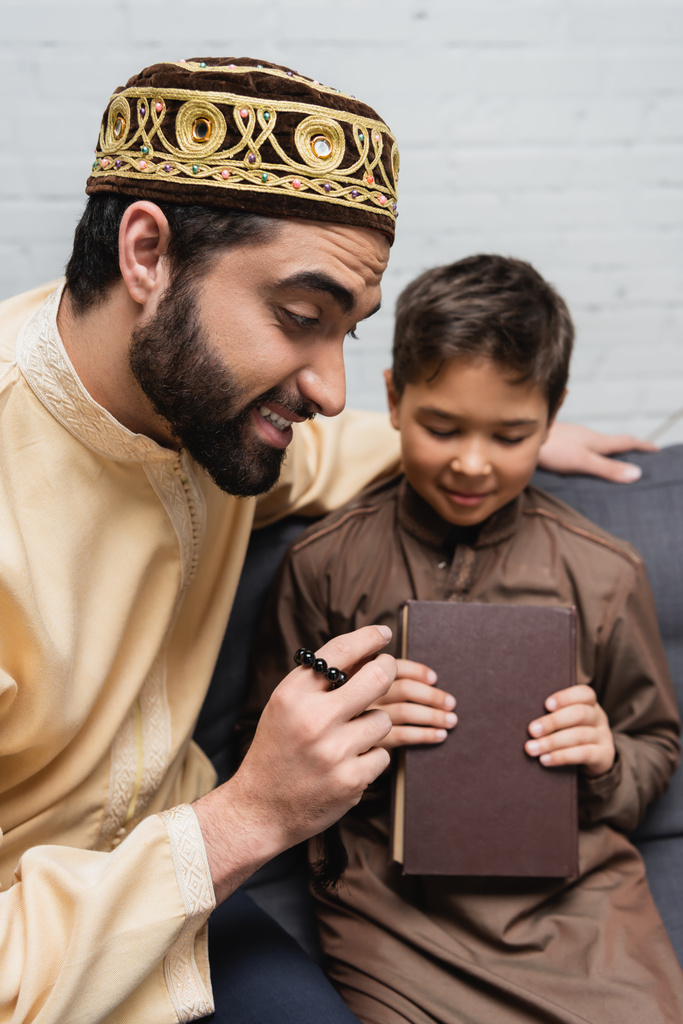 Χαμογελώντας μουσουλμάνος άνθρωπος με χάντρες προσευχή κάθεται κοντά στο γιο με το βιβλίο στον καναπέ  - Φωτογραφία, εικόνα