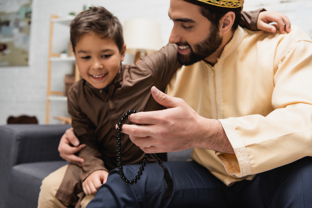 Muslim man holding prayer beads and hugging blurred son at home  - 写真・画像