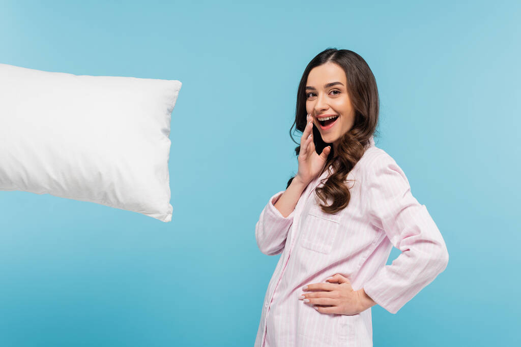 animado jovem mulher em sleepwear de pé com a mão no quadril perto levitar travesseiro isolado no azul  - Foto, Imagem