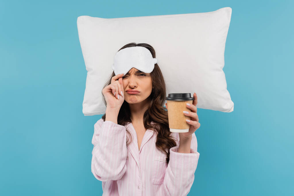 displeased woman in pajamas and sleeping mask holding paper cup with coffee to go near flying pillow isolated on blue - Fotografie, Obrázek