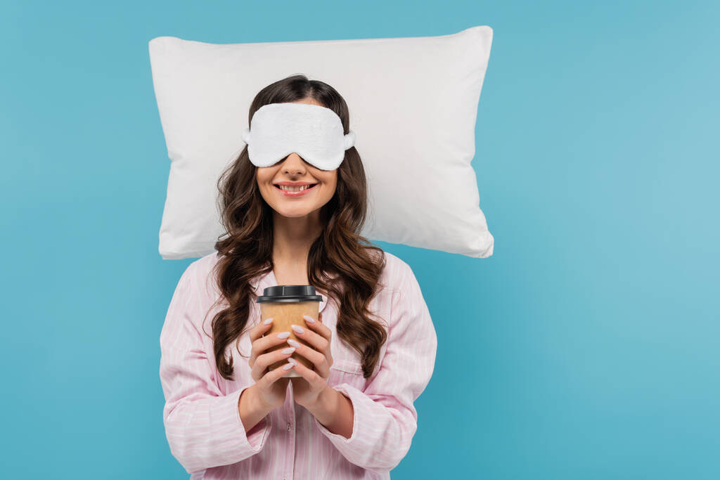 довольная женщина в пижаме и ночной маске держа бумажную чашку с кофе подойти к белой подушке изолированы на синий - Фото, изображение