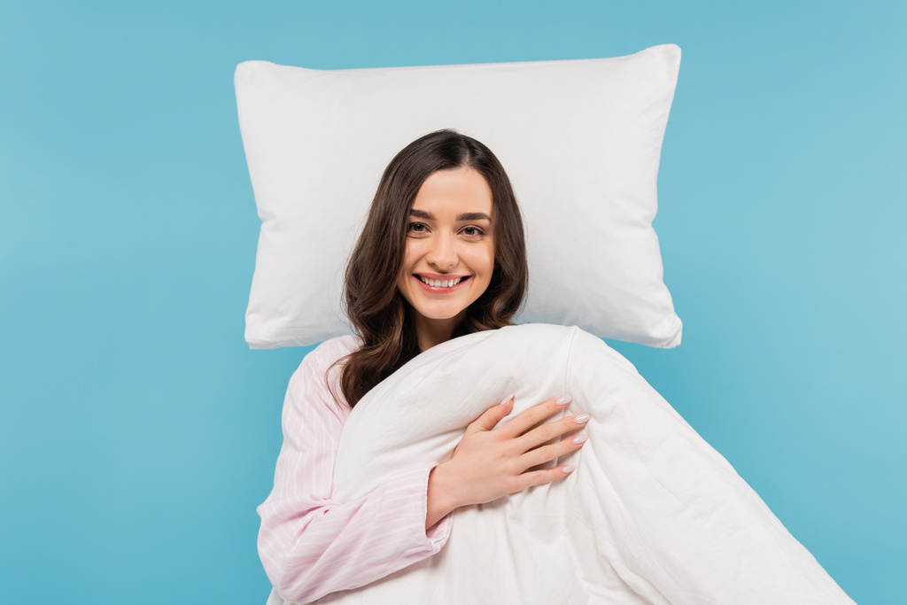 happy young woman in pajamas holding warm duvet near flying pillow isolated on blue  - Foto, afbeelding