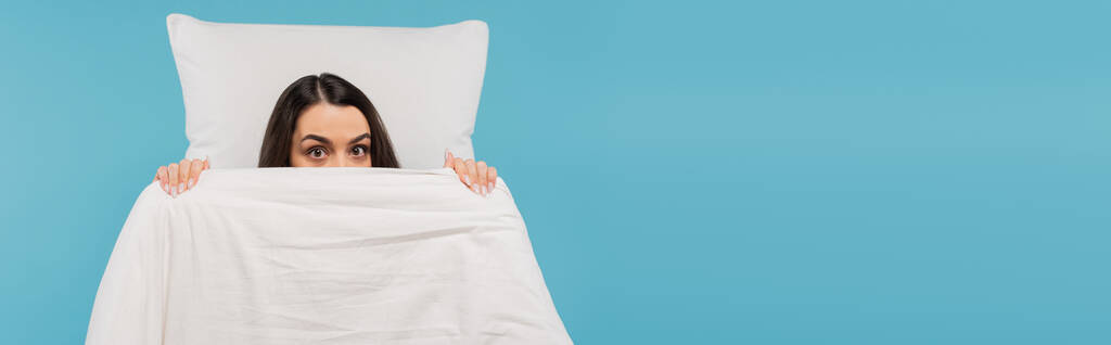 giovane donna in pigiama che copre il viso con caldo piumone vicino al cuscino isolato su blu, banner  - Foto, immagini