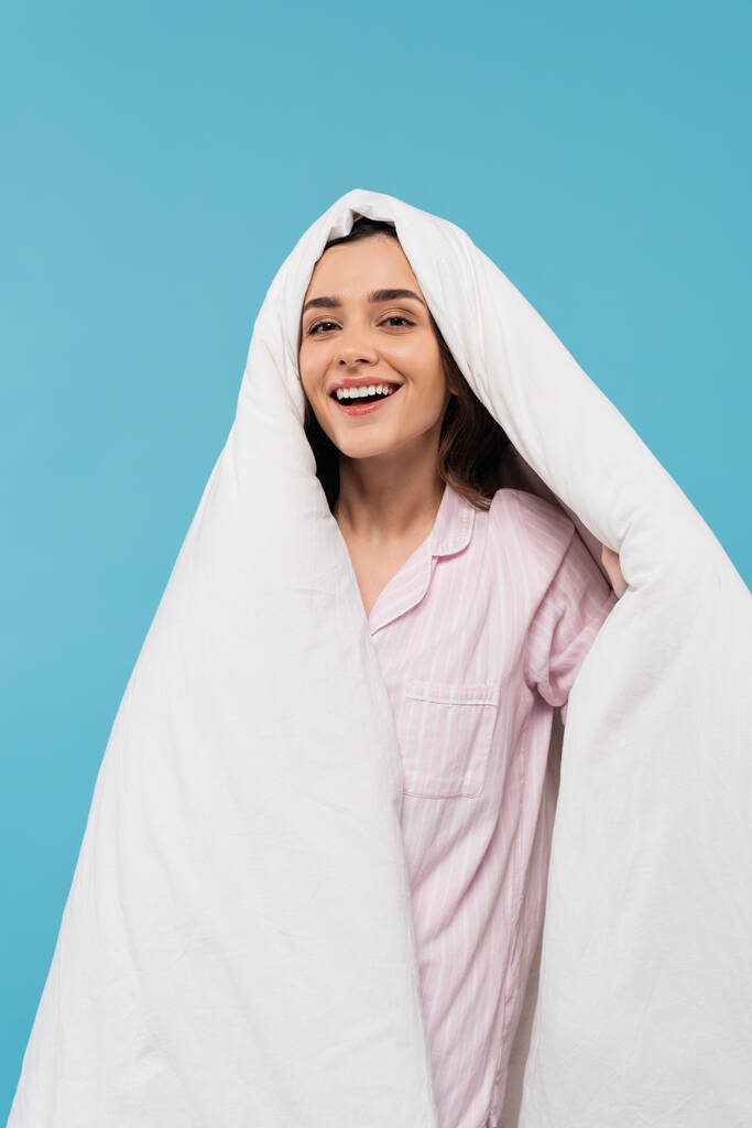 позитивная молодая женщина в пижаме покрывая голову теплым одеялом и глядя на камеру изолированы на синий  - Фото, изображение
