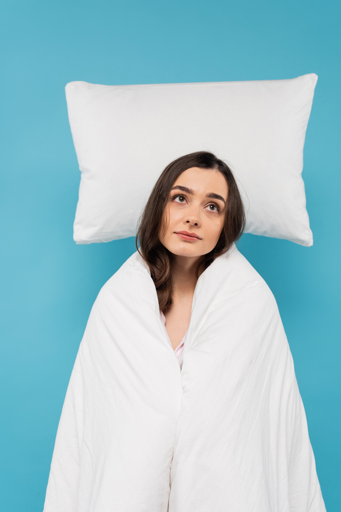 dreamy young woman covered in white duvet standing near flying white pillow on blue background  - Foto, Bild