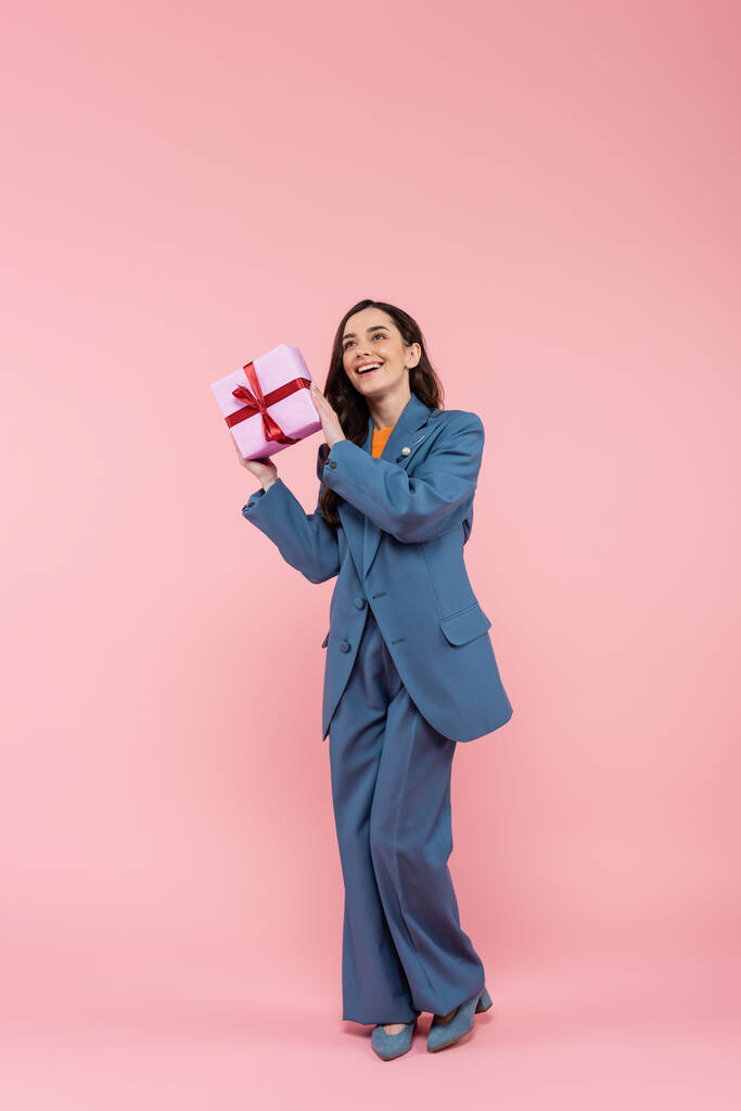full length of happy woman in blue pantsuit holding wrapped present with ribbon on pink  - Foto, Bild