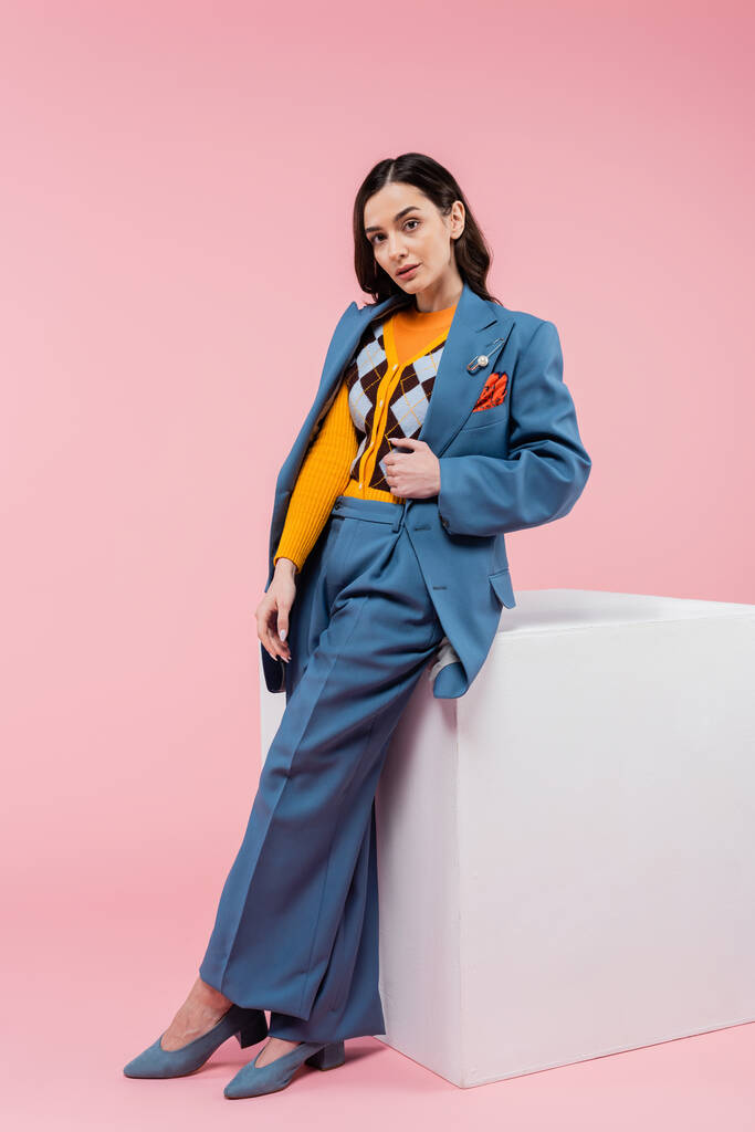 full length of brunette woman in blue suit posing near white cube and looking at camera on pink background - Foto, imagen