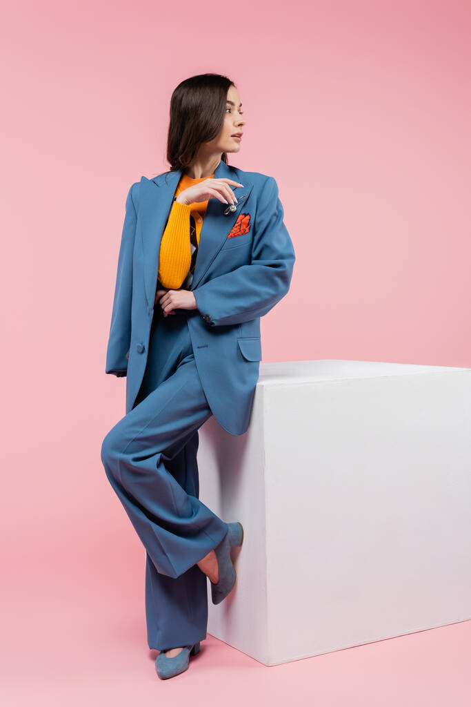 full length of brunette woman in blue suit looking away while standing near white cube on pink background - Zdjęcie, obraz