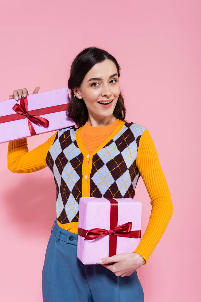 happy brunette woman in fashionable cardigan posing with presents on pink background - Photo, Image