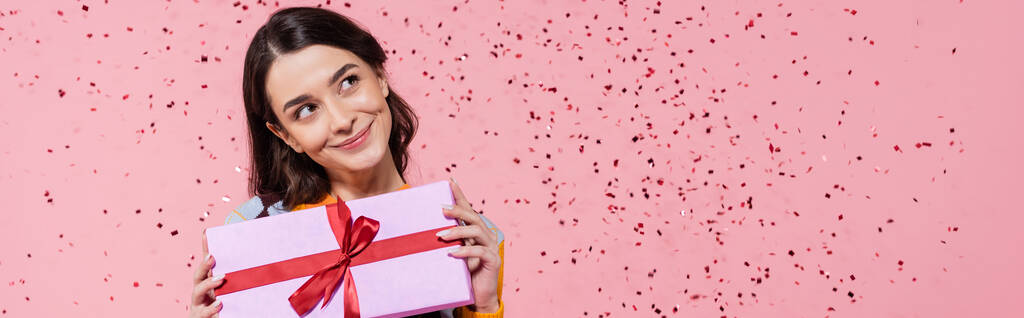 мечтательная и улыбающаяся женщина смотрит в сторону, держа подарочную коробку с красной лентой рядом конфетти на розовом фоне, баннер - Фото, изображение