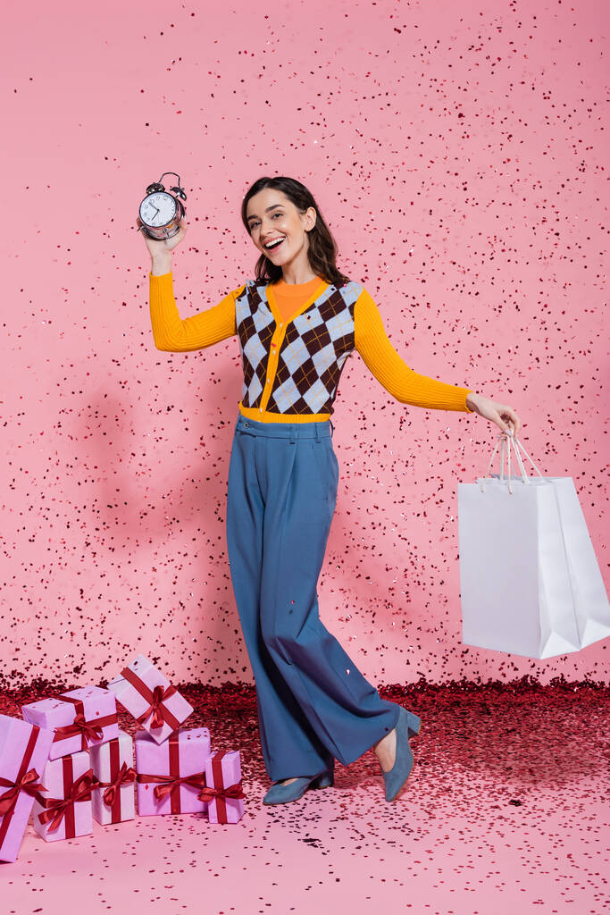 χαρούμενη και μοντέρνα γυναίκα που ποζάρει με ξυπνητήρι και τσάντες για ψώνια κοντά σε κομφετί και κουτιά δώρων σε ροζ φόντο - Φωτογραφία, εικόνα