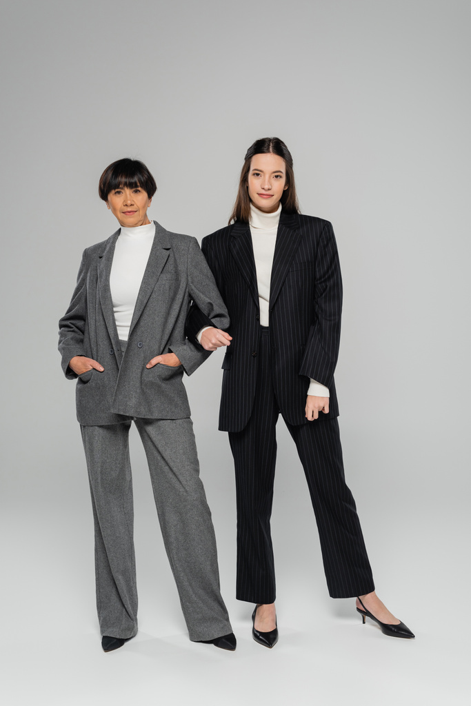 volledige lengte van positieve aziatische moeder en dochter in elegante broekpakken kijken naar camera op grijze achtergrond - Foto, afbeelding
