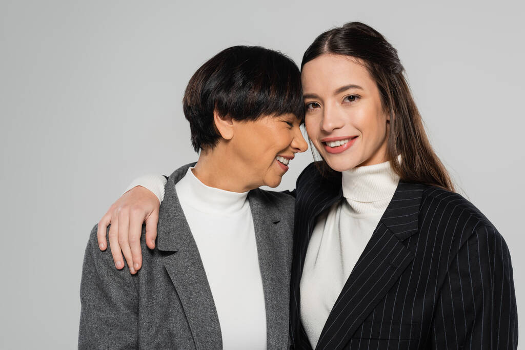 young woman in black blazer embracing happy asian mother while smiling at camera isolated on grey - Zdjęcie, obraz