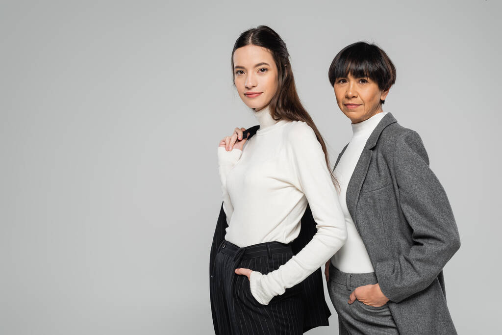 positive asian mother and daughter in formal wear posing with hands in pockets while looking at camera isolated on grey - Valokuva, kuva