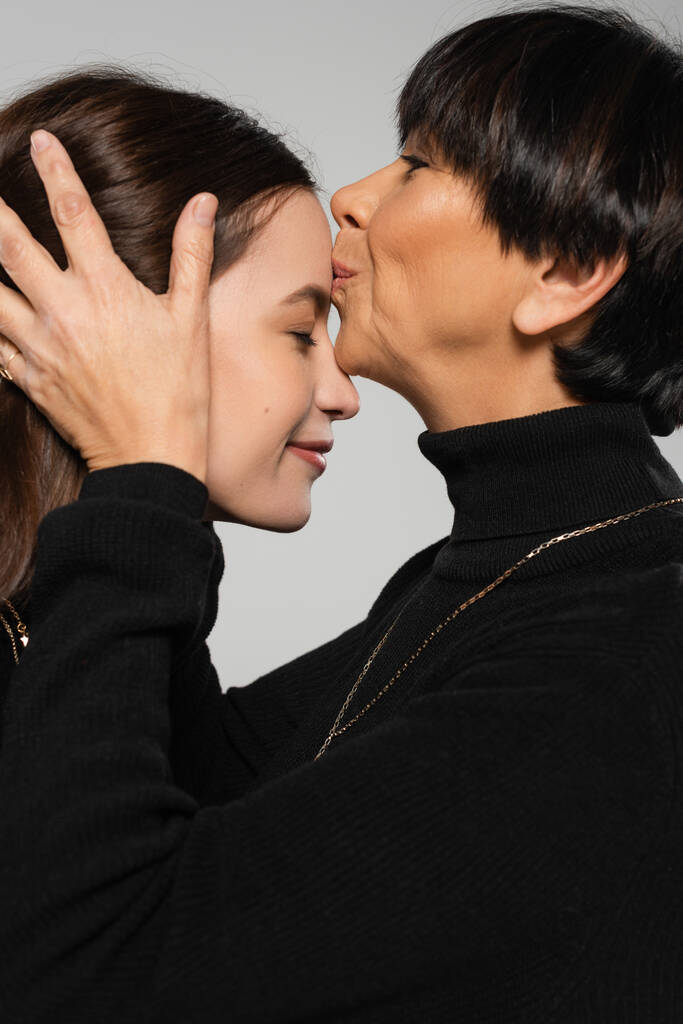 zijaanzicht van middelbare leeftijd aziatische vrouw in zwart coltrui kussen voorhoofd van jonge dochter geïsoleerd op grijs - Foto, afbeelding