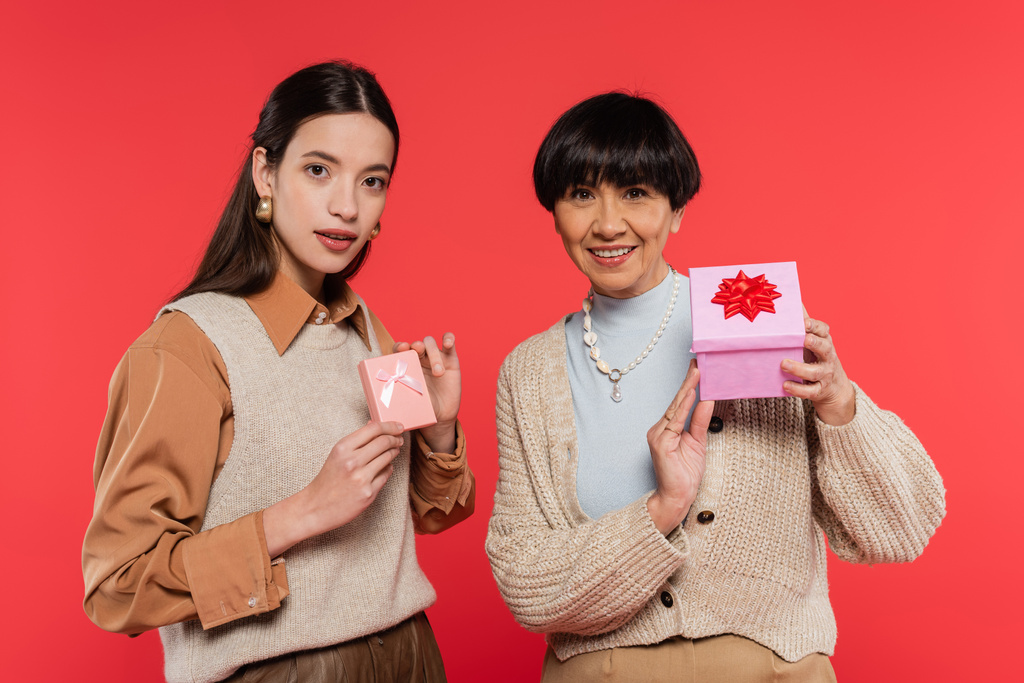 stylish asian mother and daughter showing presents and smiling at camera isolated on coral - Foto, Bild