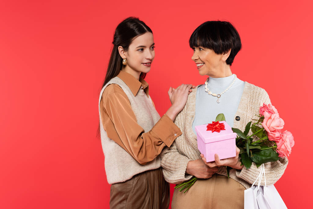 young asian daughter looking at happy middle aged mother holding flowers and gift box isolated on coral - Foto, afbeelding