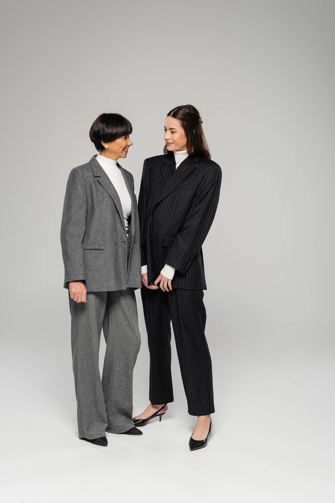 full length of cheerful asian mother and daughter in pantsuits looking at each other on grey  - Fotoğraf, Görsel