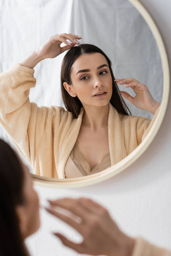 young brunette woman adjusting hair and looking at mirror in bathroom  - Fotó, kép