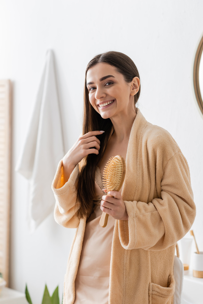 örömteli fiatal nő fürdőköpenyben kezében fa hajkefe a fürdőszobában  - Fotó, kép